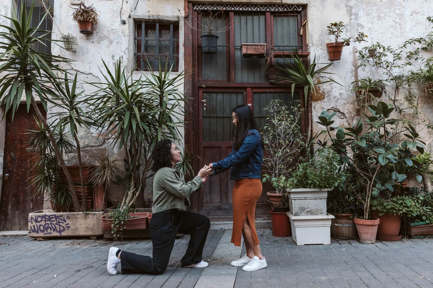 A woman is on one knee proposing to her girlfriend on a street in Barcelona with plants behind them.